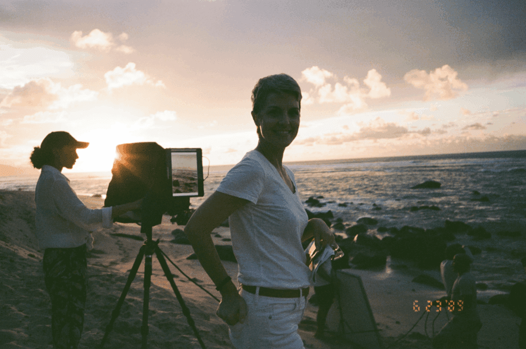 Two people stand on a rocky beach at sunset, one using a large camera on a tripod, the other posing and smiling—capturing the brand story of their memorable videoshoot; date "6.28.23" appears in the corner.