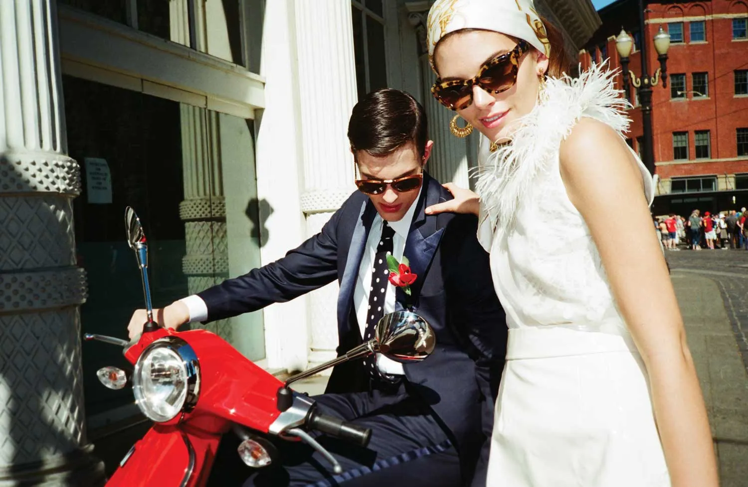 A man in a suit sits on a red scooter while a woman in a white dress and sunglasses stands beside him on a city street, capturing the essence of creative direction during an urban videoshoot.