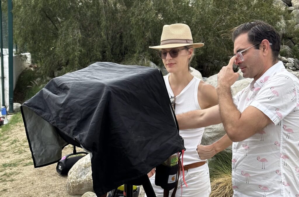 Two people stand outdoors at a videoshoot, looking at a covered monitor or camera setup; one points at the screen. Both wear sunglasses and summer clothing.