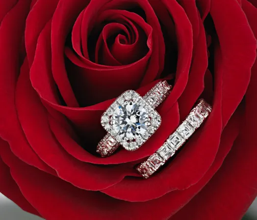 Two diamond rings rest on a red rose; guided by thoughtful visual direction, one dazzles with a large round diamond in a square halo, while the other shines with a row of square-cut diamonds.