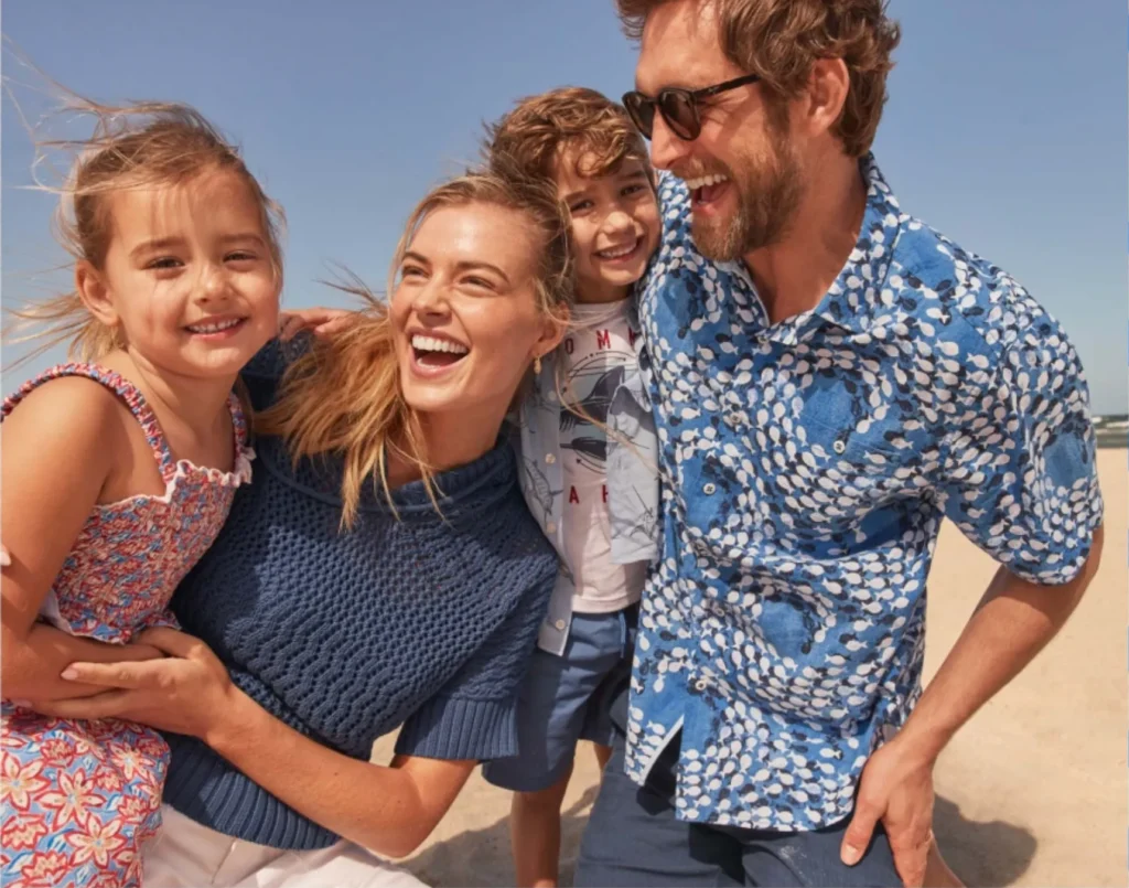 A smiling family of four, two adults and two children, stand close together on a sandy beach under a clear blue sky, perfectly posed for a photoshoot.