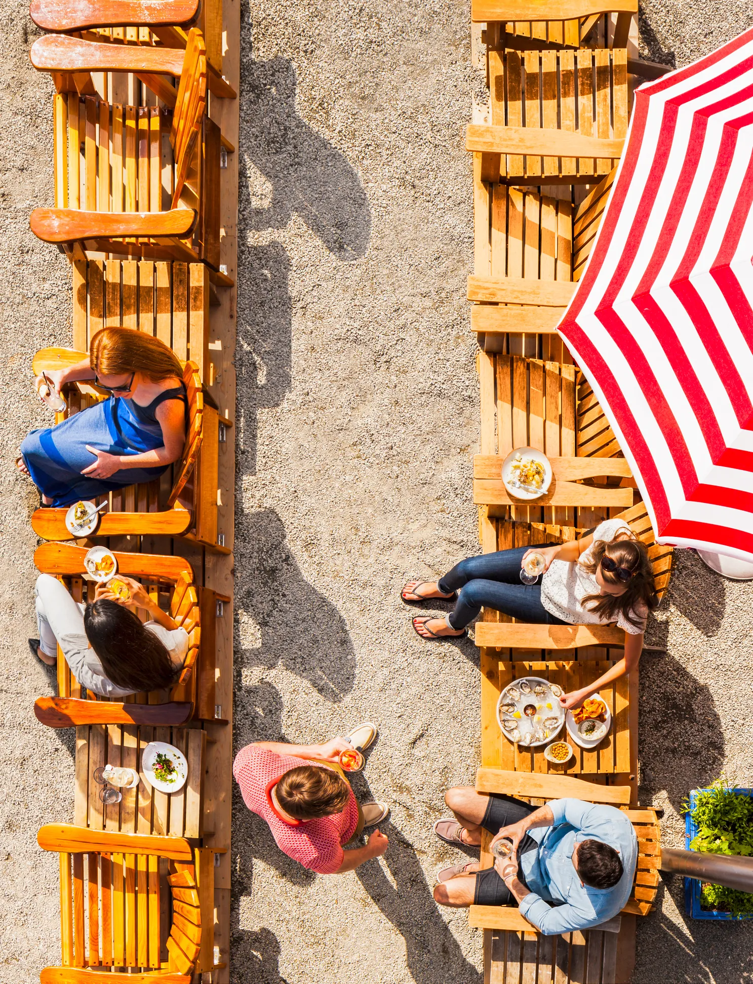 Overhead view of people sitting in wooden chairs around tables with food and drinks, next to a red and white striped umbrella on a gravel surface, showcasing creative direction ideal for a vibrant videoshoot.