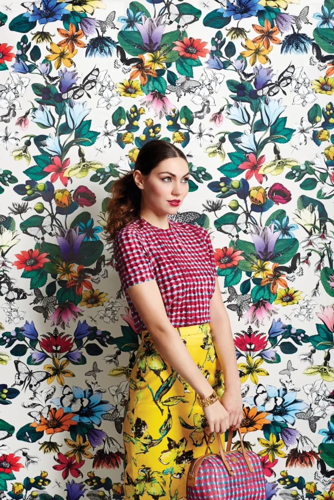 A woman stands against a colorful floral and butterfly wallpaper in a vibrant photoshoot, wearing a red checkered top and yellow skirt with a floral pattern, holding a matching red checkered bag.