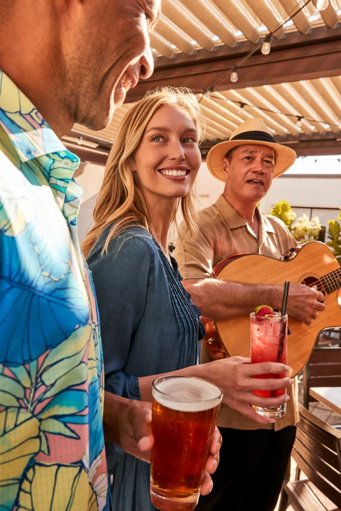 Three people stand outdoors during a creative direction photoshoot; one woman holds a cocktail and smiles at the camera, one man holds a beer, and another man plays guitar.
