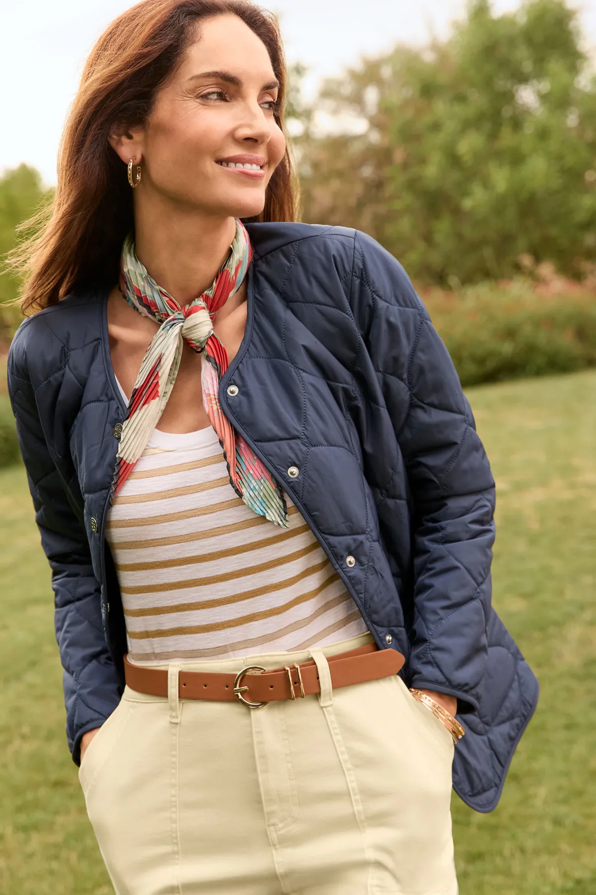 Woman outdoors in a navy quilted jacket, striped top, beige pants with a brown belt, and a scarf tied around her neck, standing on grass with trees behind her—capturing thoughtful visual direction for an inspiring photoshoot.