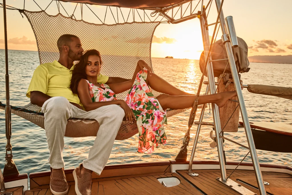 A couple relaxes together on a hammock aboard a sailboat at sunset, with calm water and a clear sky in the background—an idyllic setting perfect for a videoshoot guided by a creative director’s visual direction.